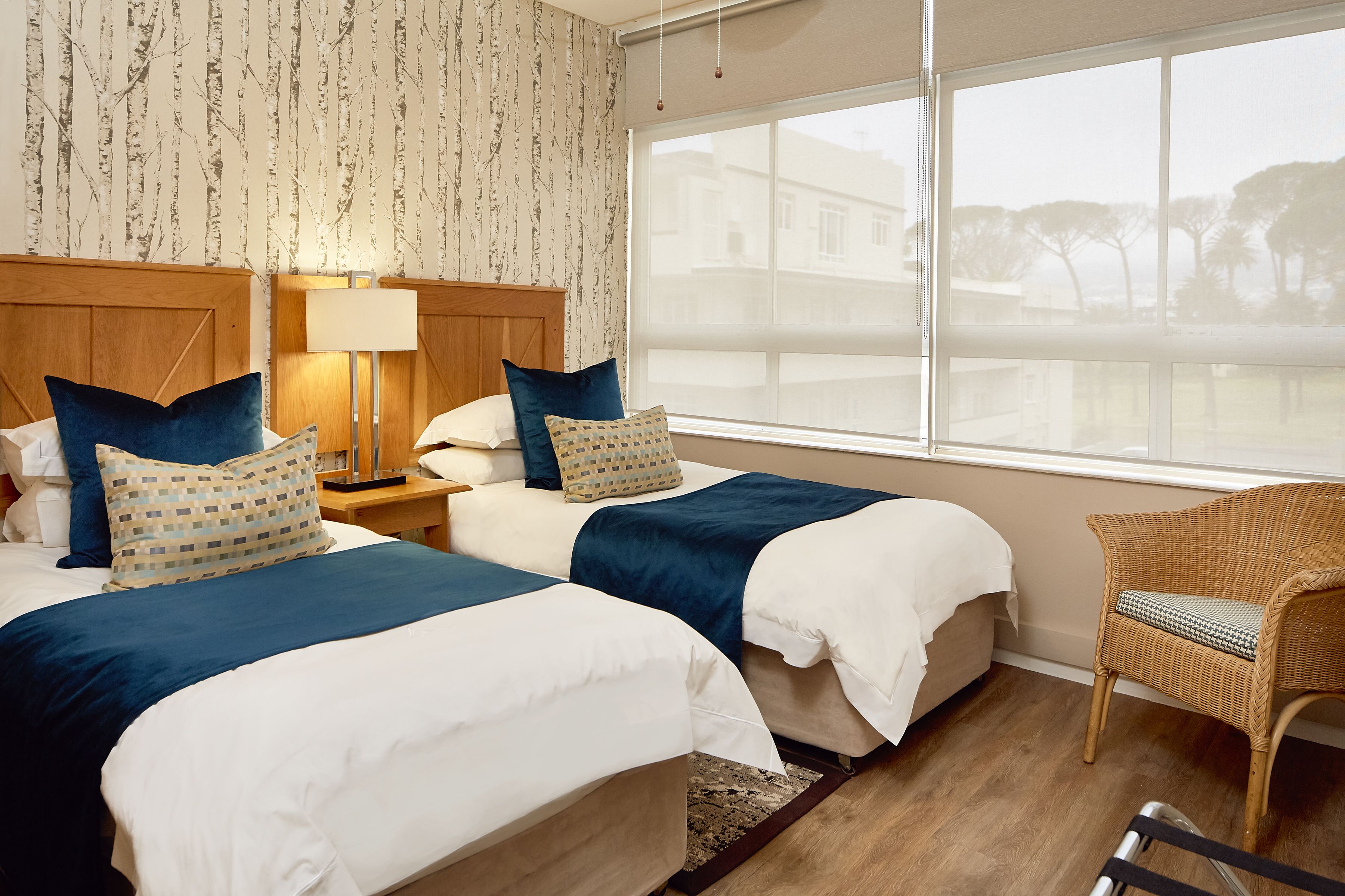 A hotel room with two beds, a wooden headboard, a wicker chair, and a large window with a view of buildings and trees.
