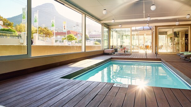 Una piscina interior con plataforma de madera, ventanas amplias que ofrecen vistas a una montaña y edificios, y una puerta de cristal que da a un área exterior con mobiliario.