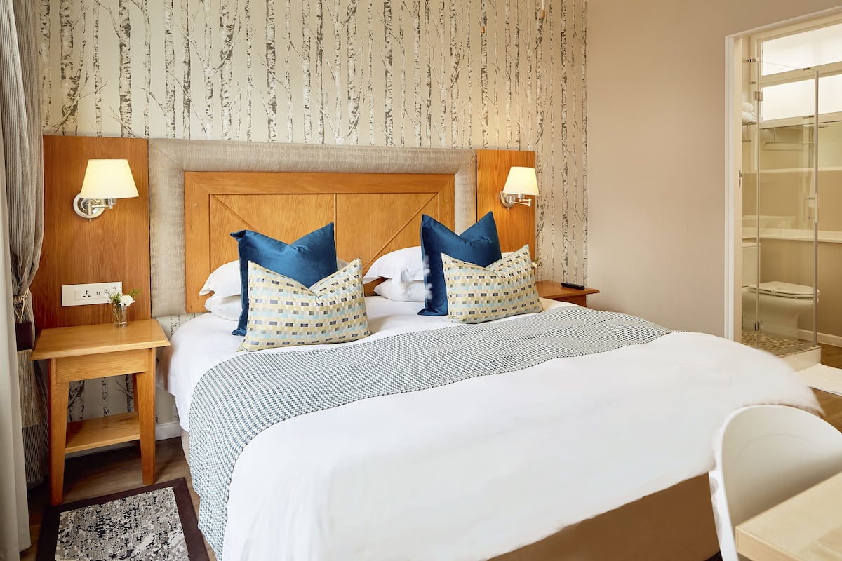 Une chambre d’hôtel avec un grand lit, deux tables de chevet, une tête de lit en bois et une salle de bain visible à travers une porte ouverte.