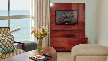 Een hotelkamer met een groot raam met uitzicht op de zee, een houten tv-meubel met een flatscreen-televisie, een eettafel met een bloemenvaas en een bank.