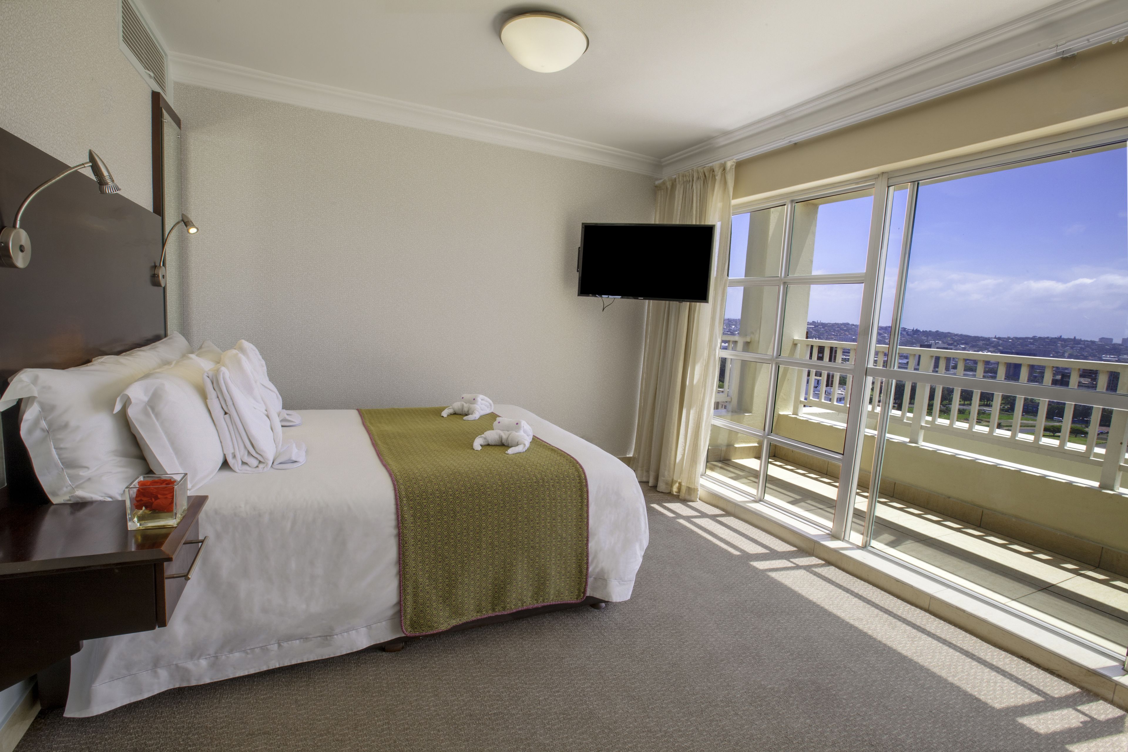 A hotel room with a large bed, a flat-screen TV, a bedside table with a glass, and a view of the city through large windows.