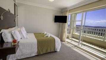 A hotel room with a large bed, a flat-screen TV, a bedside table with a glass, and a view of the city through large windows.