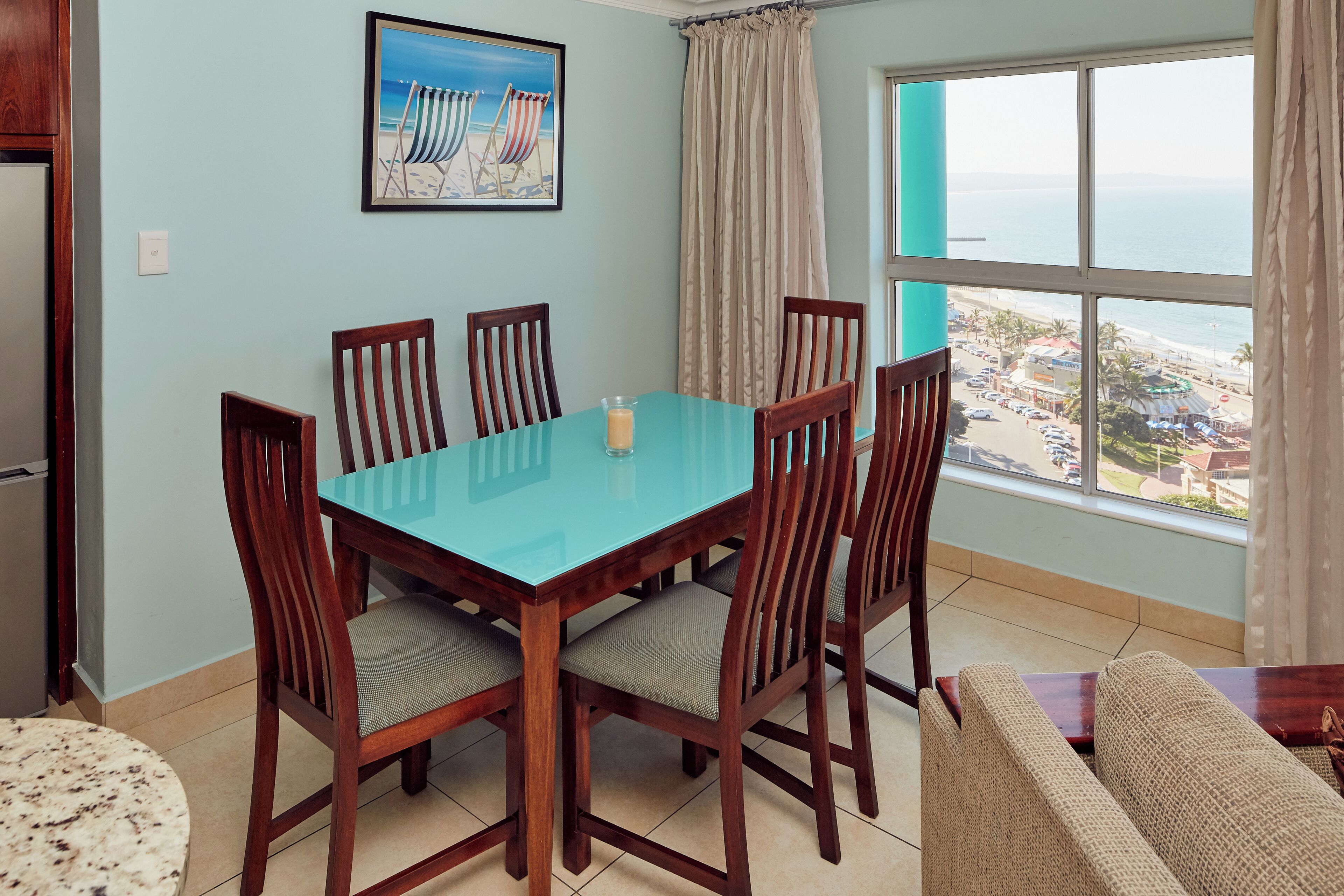 Un comedor con mesa de vidrio y sillas de madera, un cuadro de una playa en la pared, y vista a una playa y un pueblo a través de un ventanal grande.