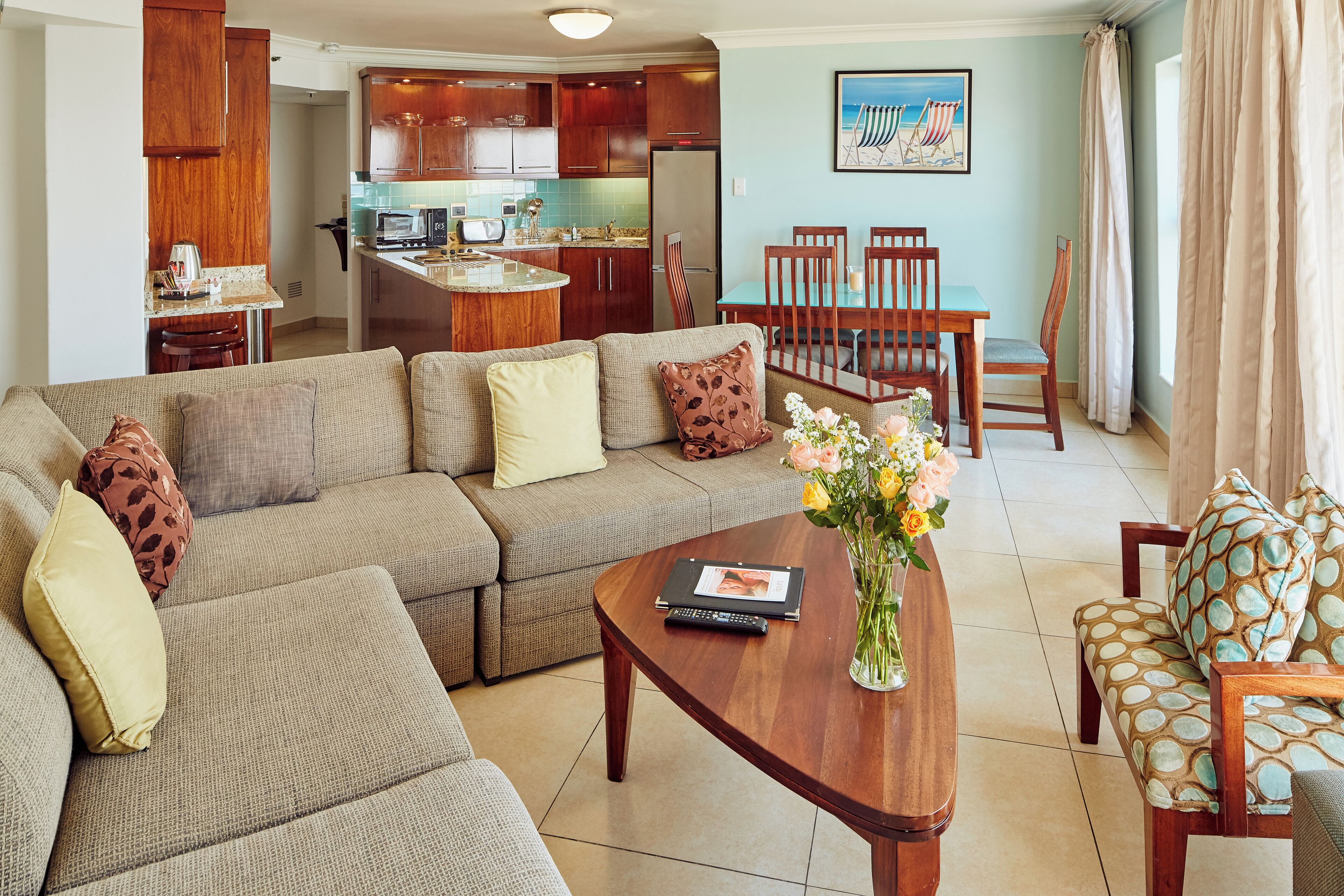 A living room with a sofa, coffee table, and chairs. There is a kitchen in the background with wooden cabinets and a dining table.