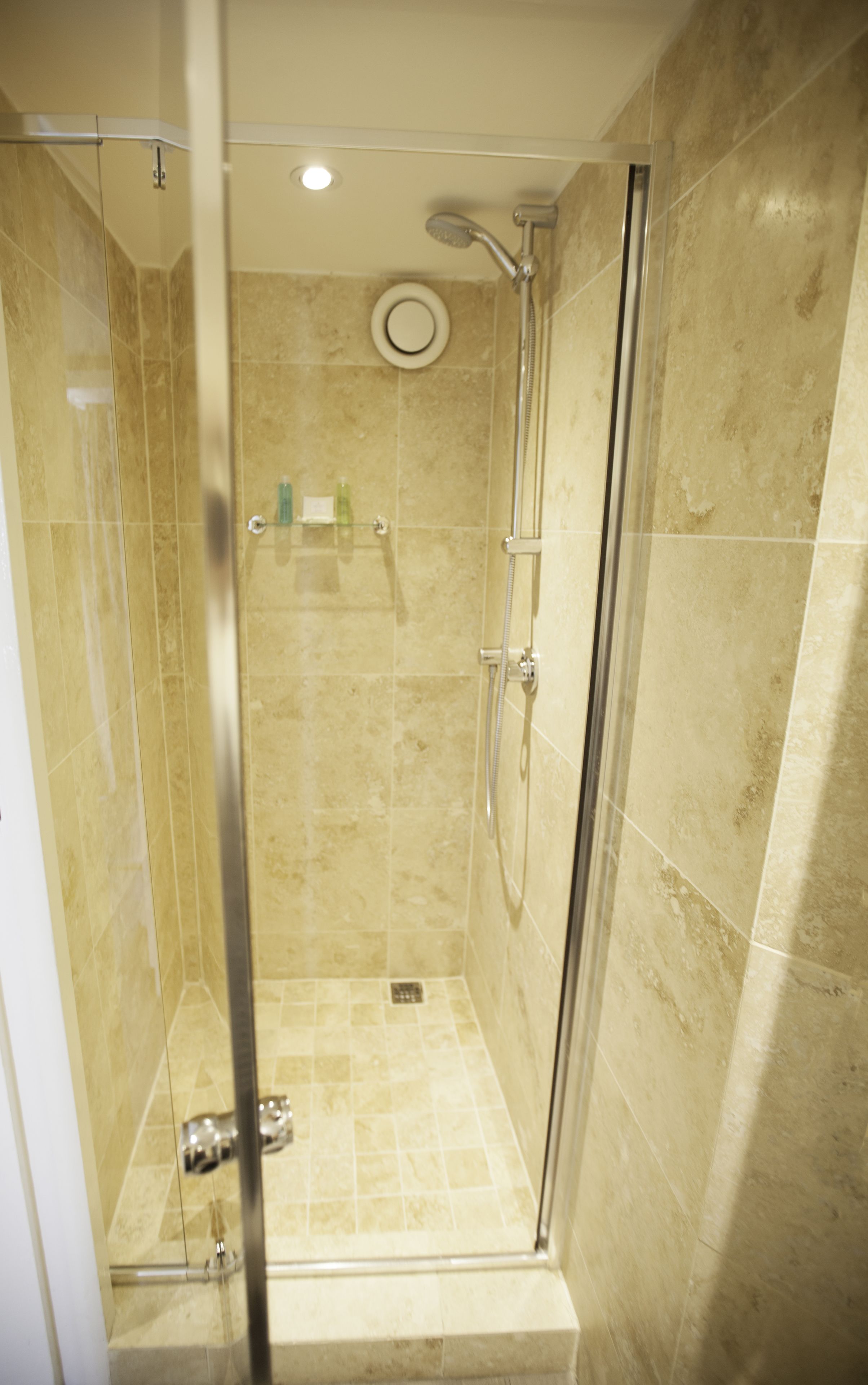 A glass-enclosed shower stall with a handheld showerhead, a wall-mounted control panel, and a small shelf with toiletries.