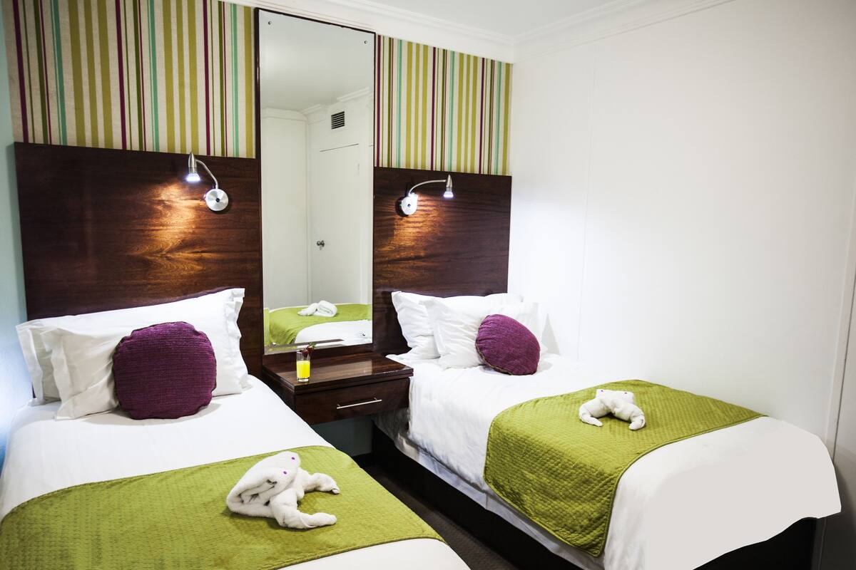 A hotel room with two beds, each with a green blanket and a white pillow. There are two bedside tables with lamps. The room has a striped wallpaper and a large mirror above the beds.
