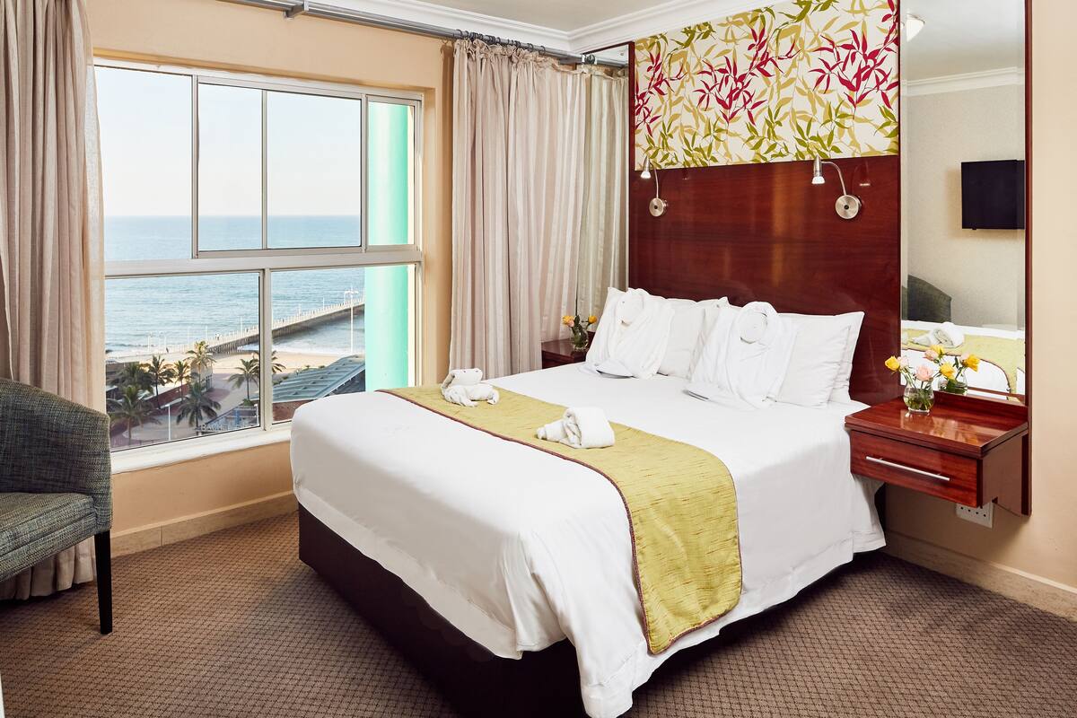 A hotel room with a large bed, a view of the ocean, a wooden headboard, a bedside table with a lamp, and a chair.