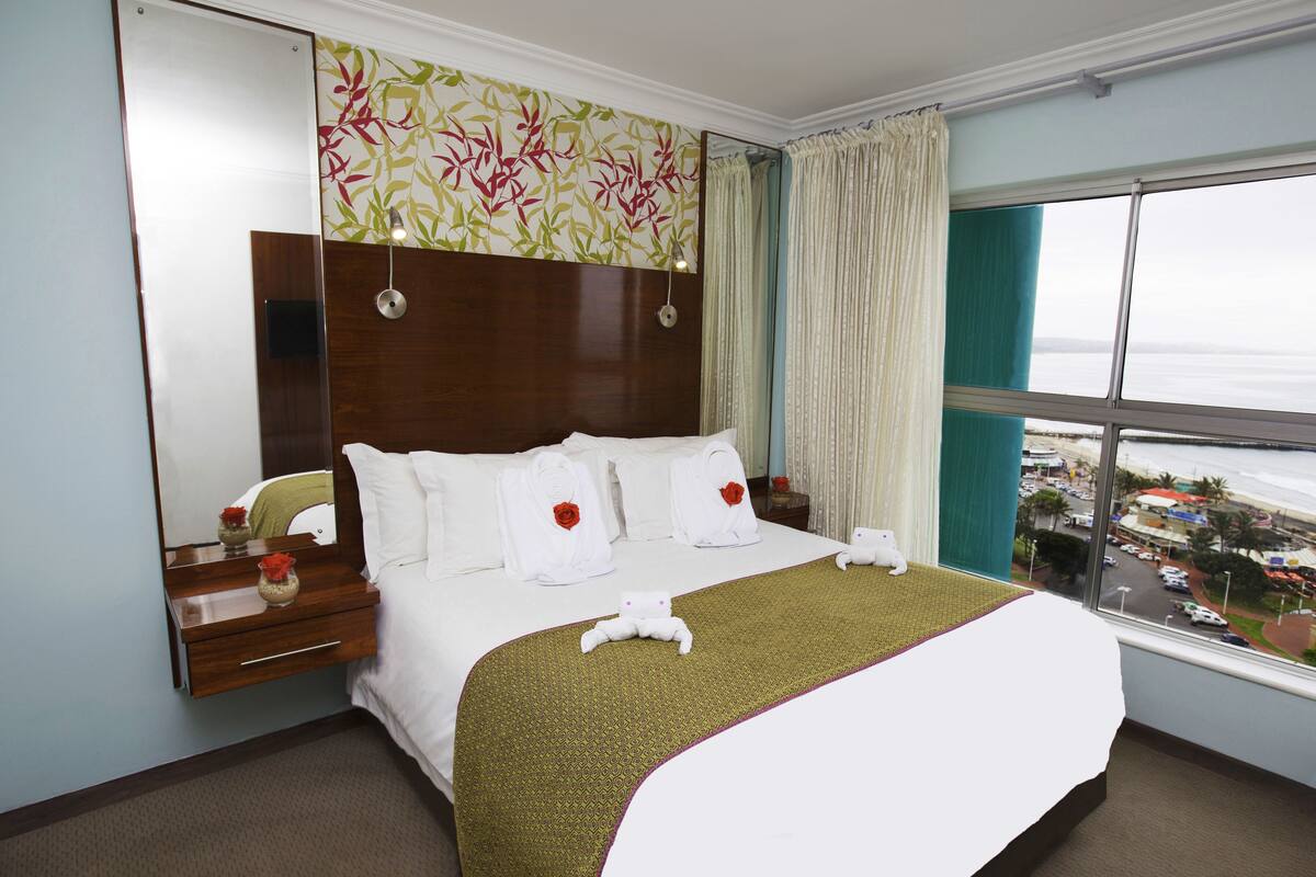 A hotel room with a large bed, a nightstand, a mirror, and a window with a city view.