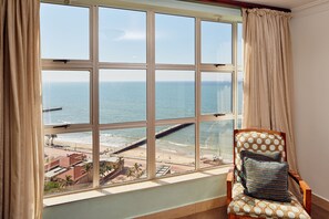 A room with a large window overlooking a beach, a wooden chair with a patterned cushion, and a view of the ocean. - First Group The Palace All-Suite (Durban)