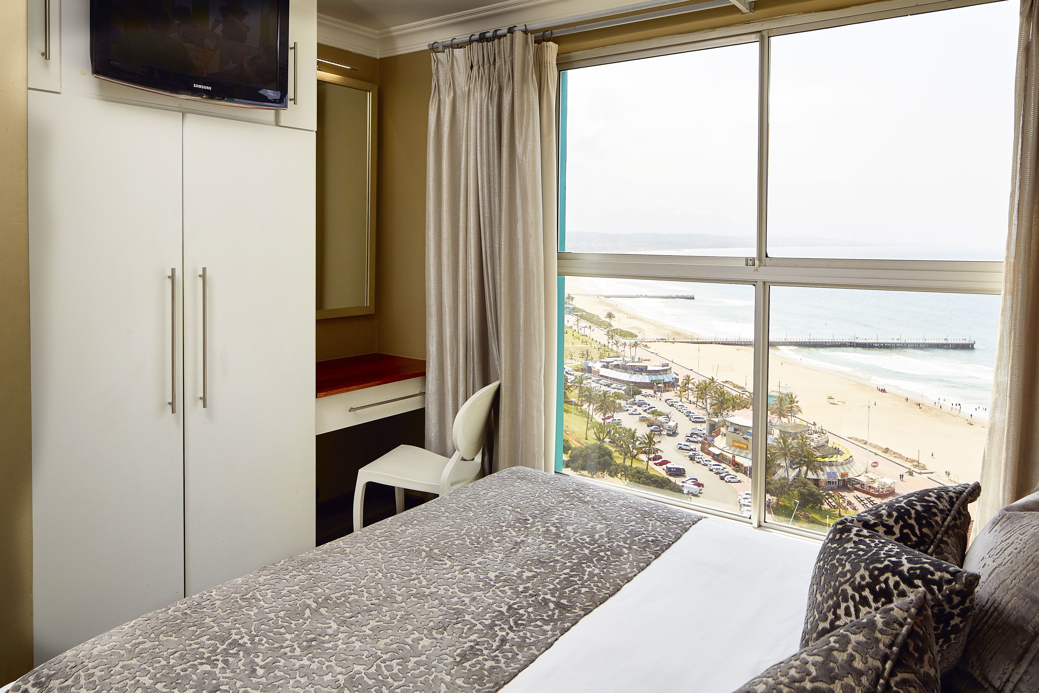 A hotel room with a bed, a desk, a chair, and a large window offering a view of the beach and ocean.