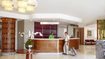 A modern hotel reception area with a wooden reception desk, a large potted orchid, and a chandelier.