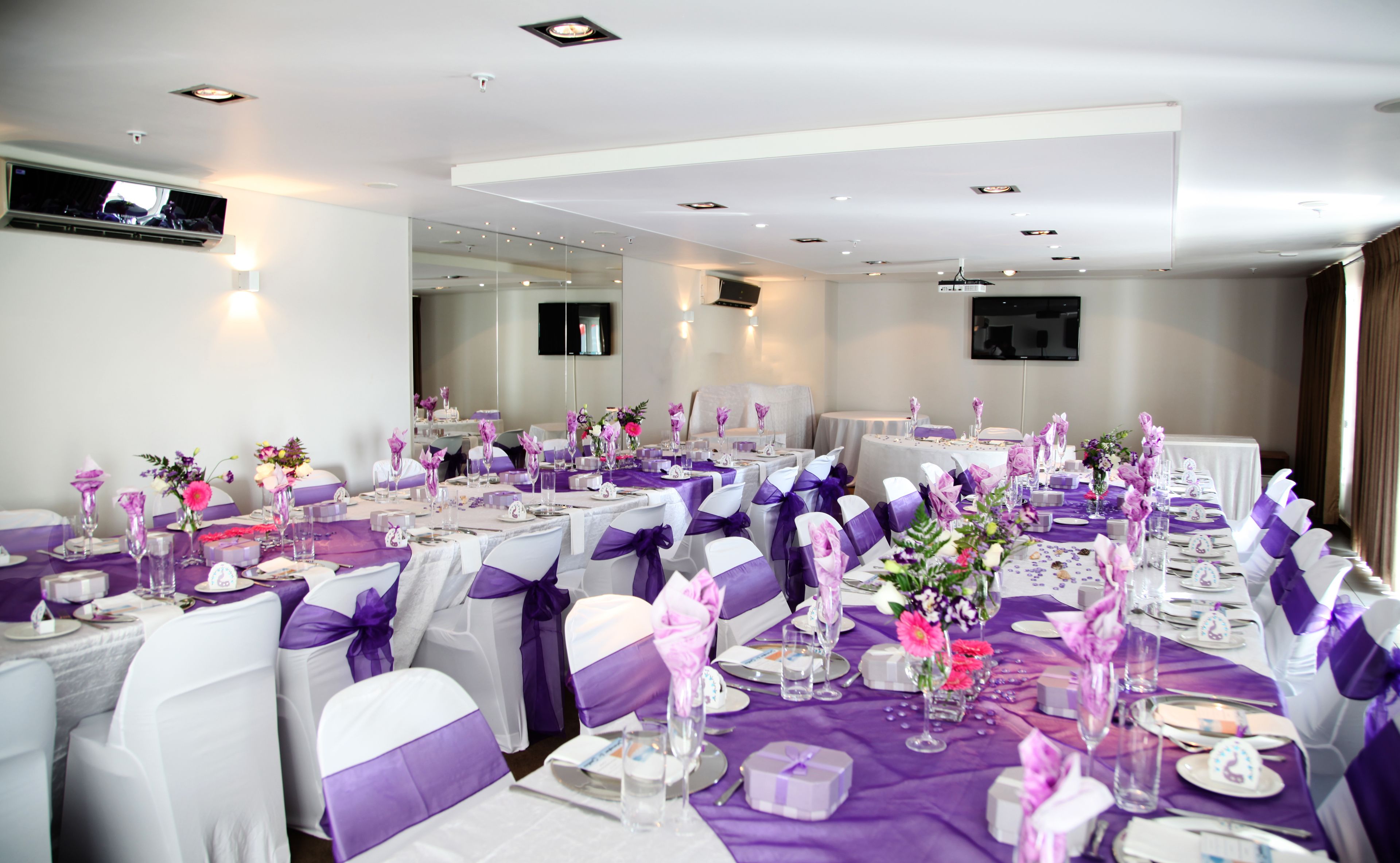 A banquet hall with tables set for a formal event, featuring purple tablecloths and white chairs with purple sashes.