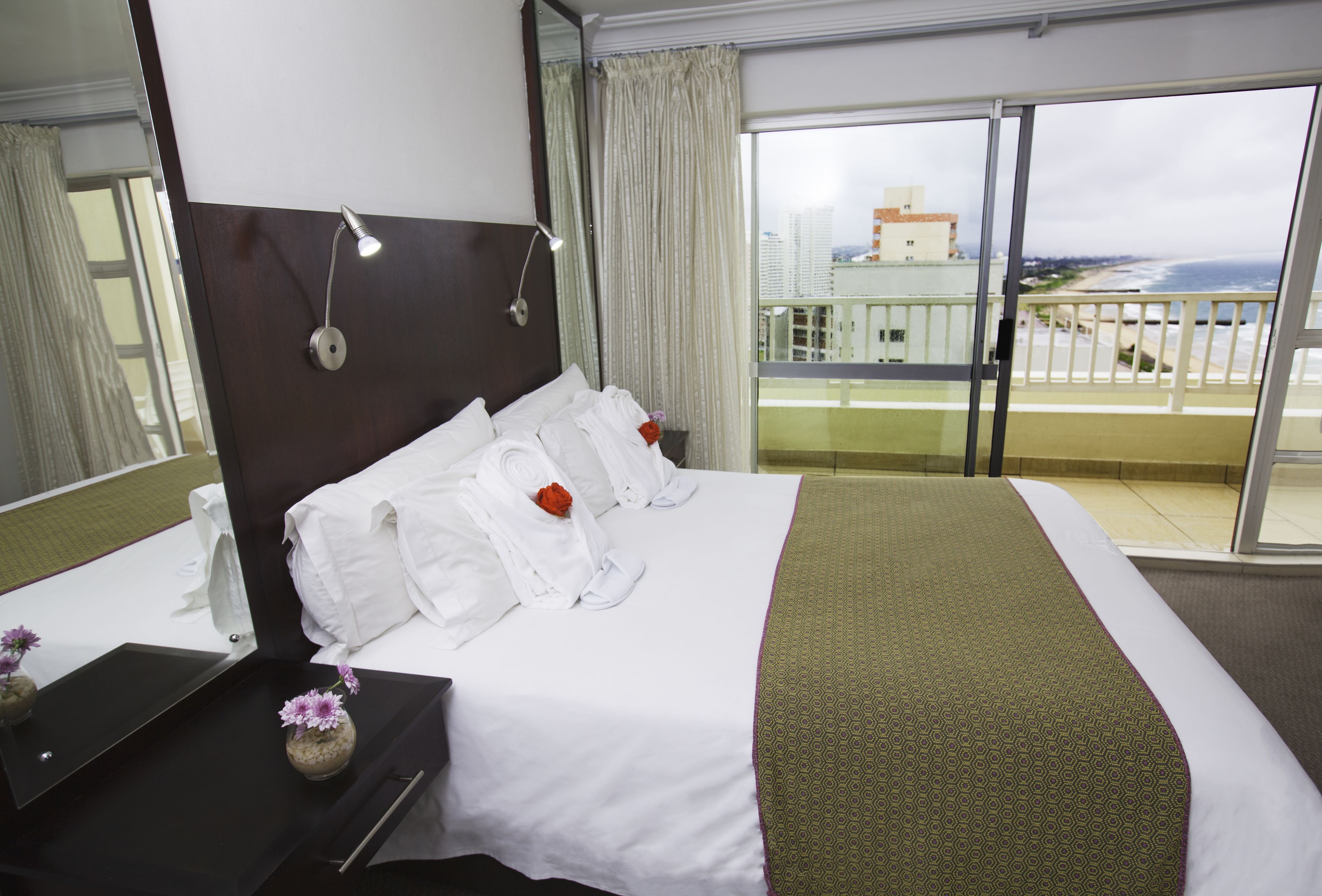 A hotel room with a large bed, a nightstand, a mirror, and a view of the ocean through a balcony.