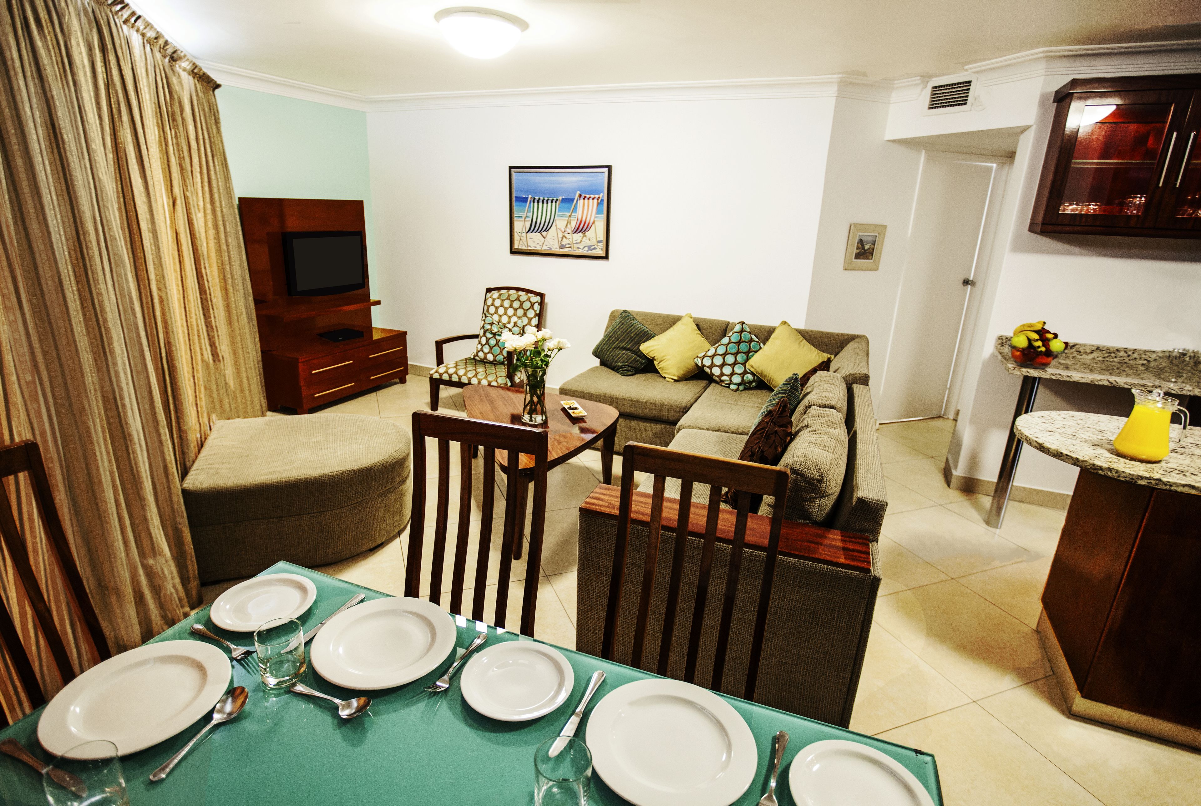 Un comedor con una mesa dispuesta para cuatro personas, un televisor, un sofá y una zona de cocina.