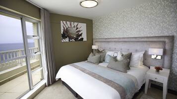 A hotel room with a large bed, a nightstand with lamps, a painting on the wall, and a view of the ocean through a sliding door.
