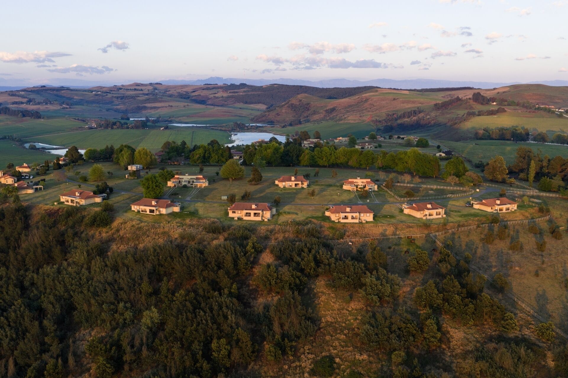 An aerial view of a residential area with multiple houses, a lake, and rolling hills in the background.