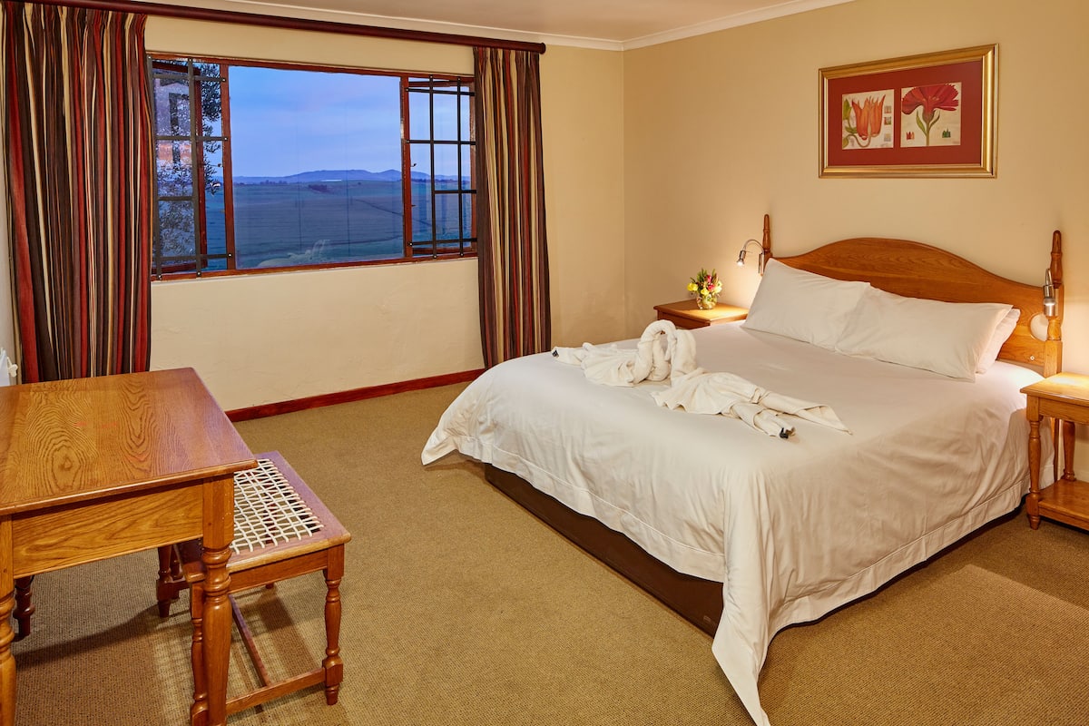 A hotel room with a large bed, a wooden nightstand with a lamp, a small table, and a window with curtains.