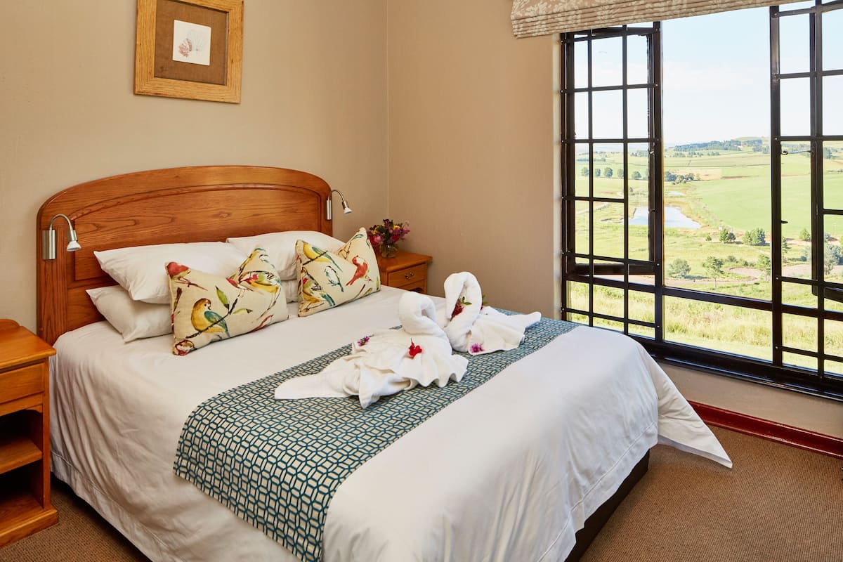 A neatly made bed with a patterned bedspread, a wooden headboard, and a view of a green landscape through a large window.