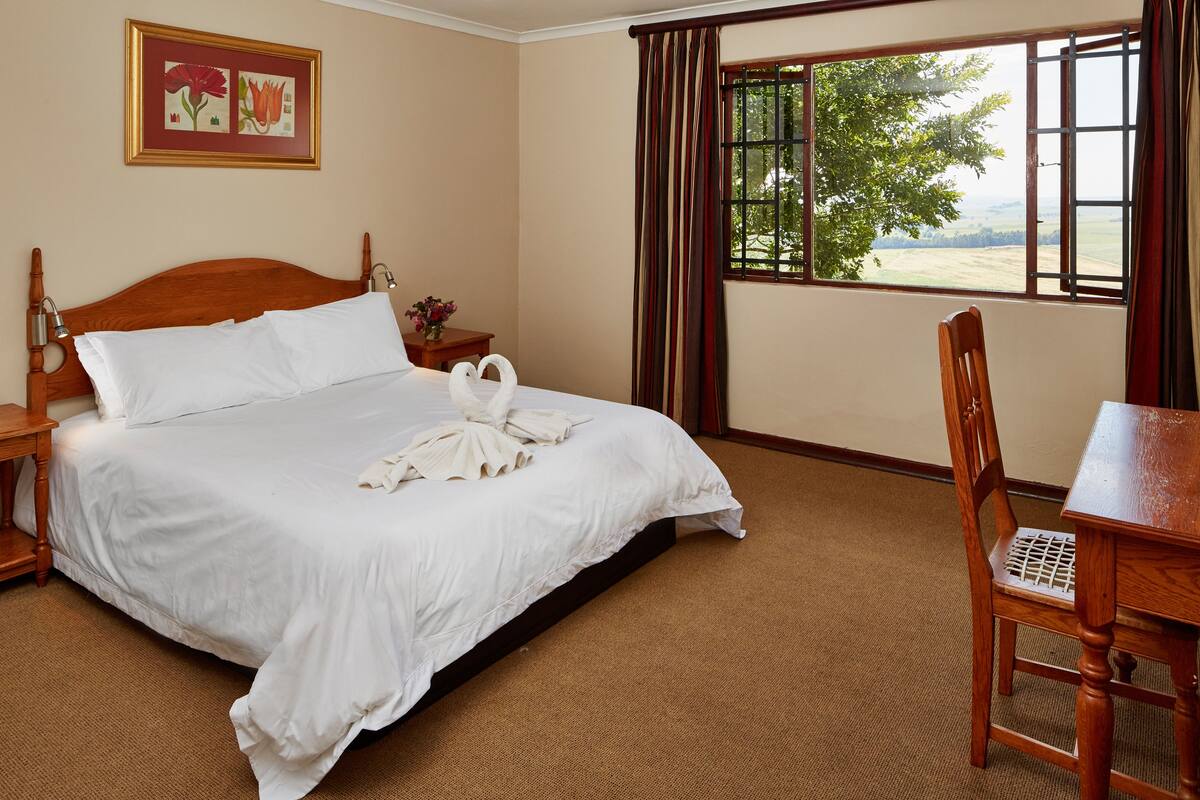 A neatly made bed with white linens and two swan-shaped towels, a wooden nightstand with a vase of flowers, a wooden chair, and a window with a view of greenery.