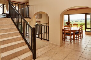 A staircase with black railings leading to a dining area with a table and chairs, and a view of the countryside through large windows. - First Group Midlands Saddle and Trout (Mooirivier)