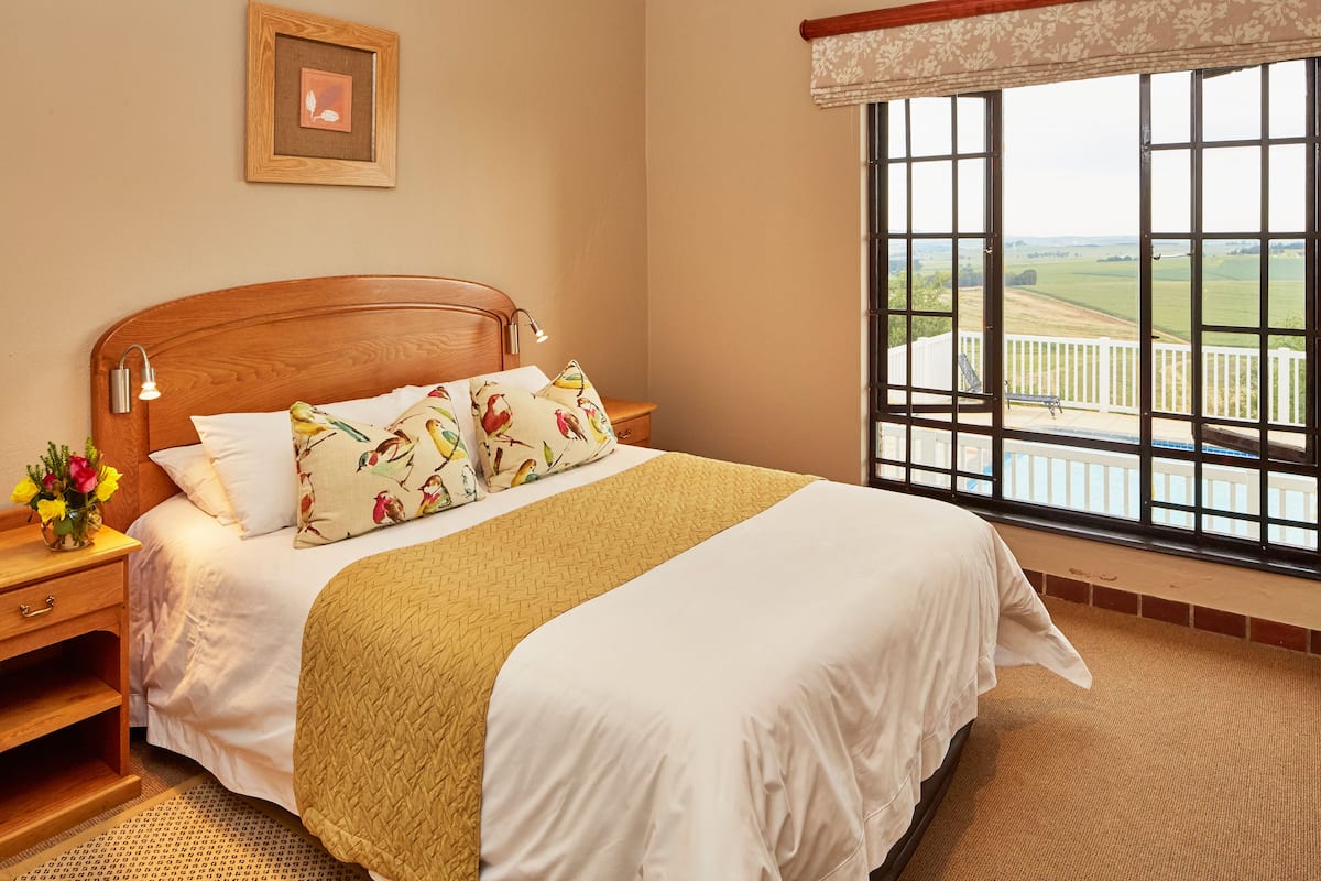 A neatly made bed with a wooden headboard, a bedside table with a lamp, and a view of a balcony with a railing.