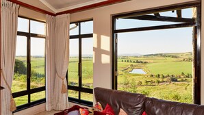 A room with a large window offering a view of a green landscape, a brown leather sofa, and a small table with a vase. - First Group Midlands Saddle and Trout (Mooirivier)