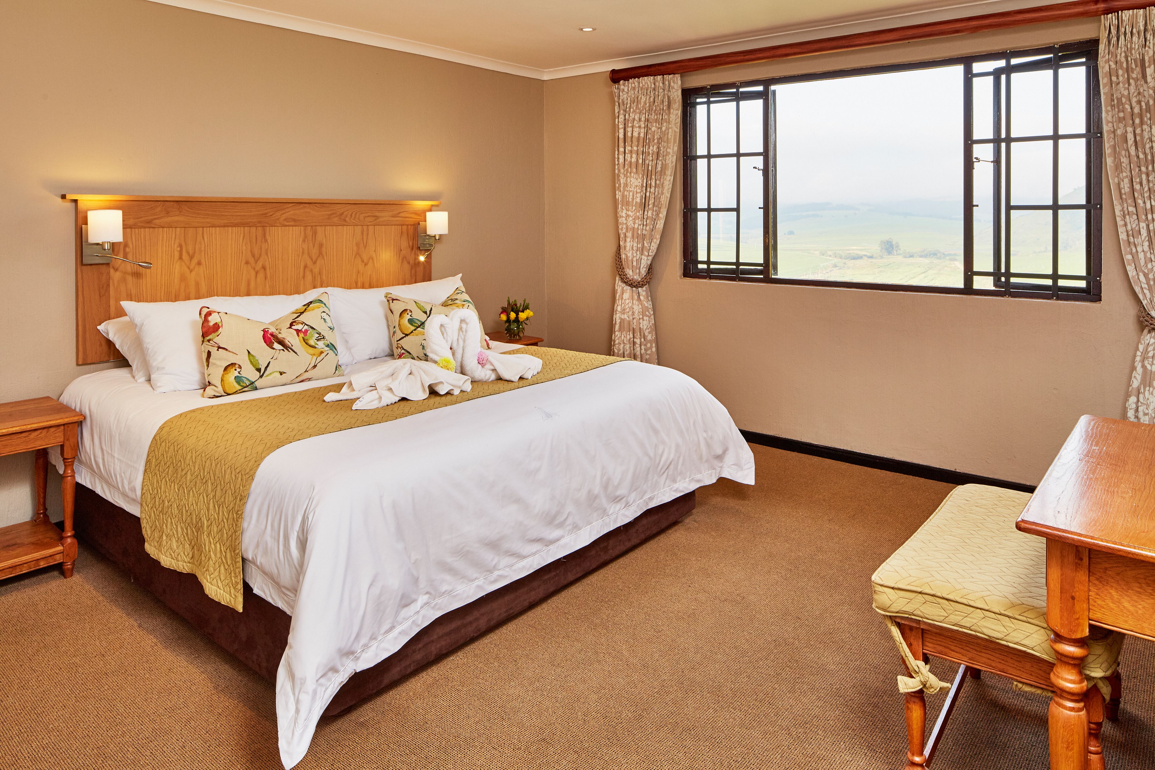 A neatly made bed with white linens and a yellow throw, a wooden headboard, and a view of the countryside through a large window.