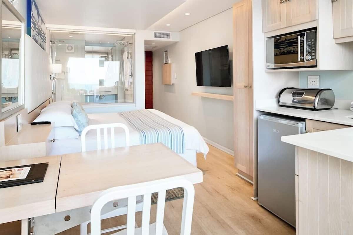 A compact hotel room with a bed, a small dining table, a kitchenette with a microwave, and a TV mounted on the wall.