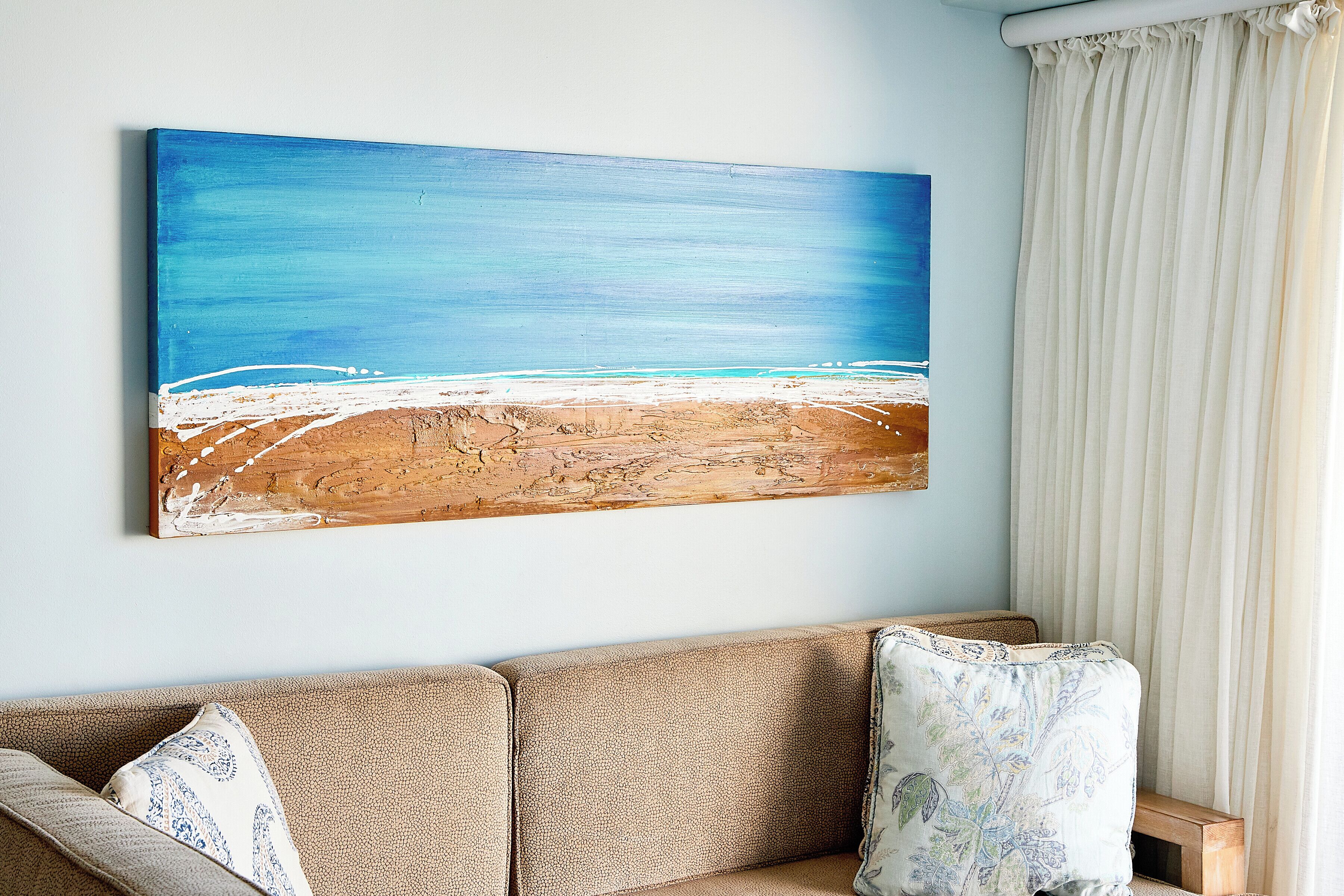 A room with a beige sofa, a blue and white painting of a beach, and a white curtain.
