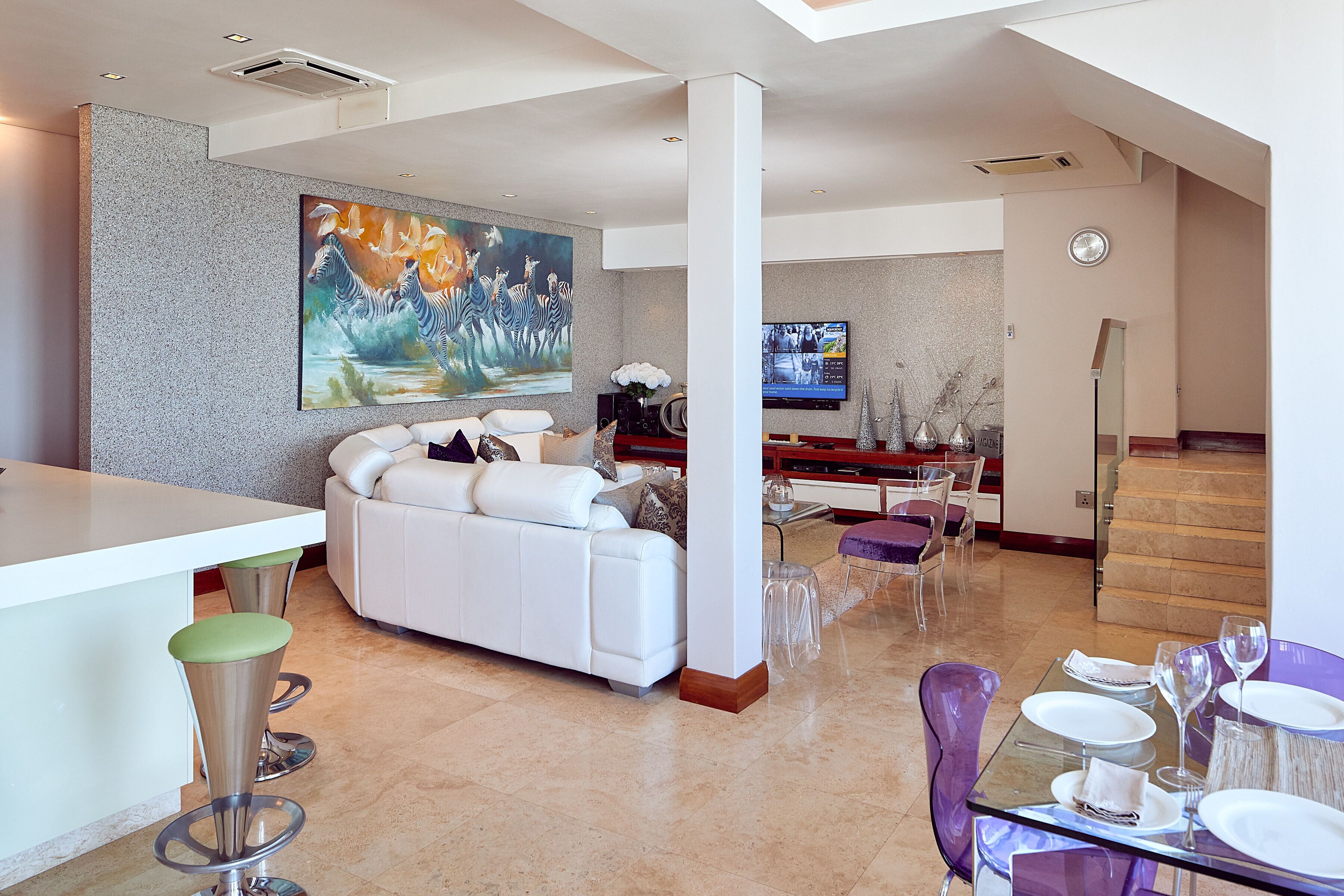 A modern living room with a white sofa, a large painting, a TV, and a dining area with a glass table and purple chairs.
