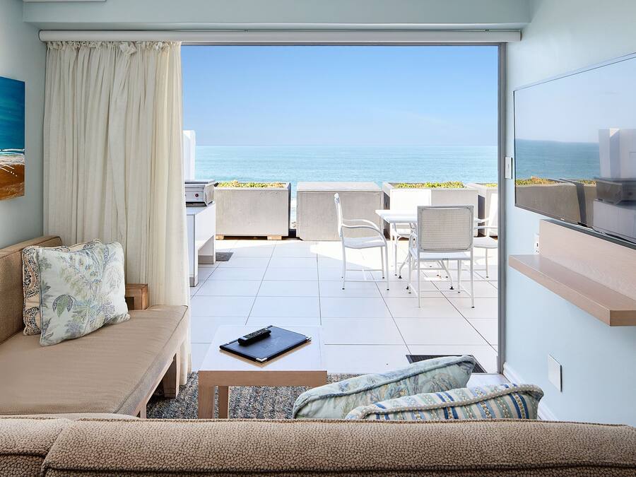 A modern living room with a sofa, a coffee table, and a TV. There is a view of the ocean through a glass door.