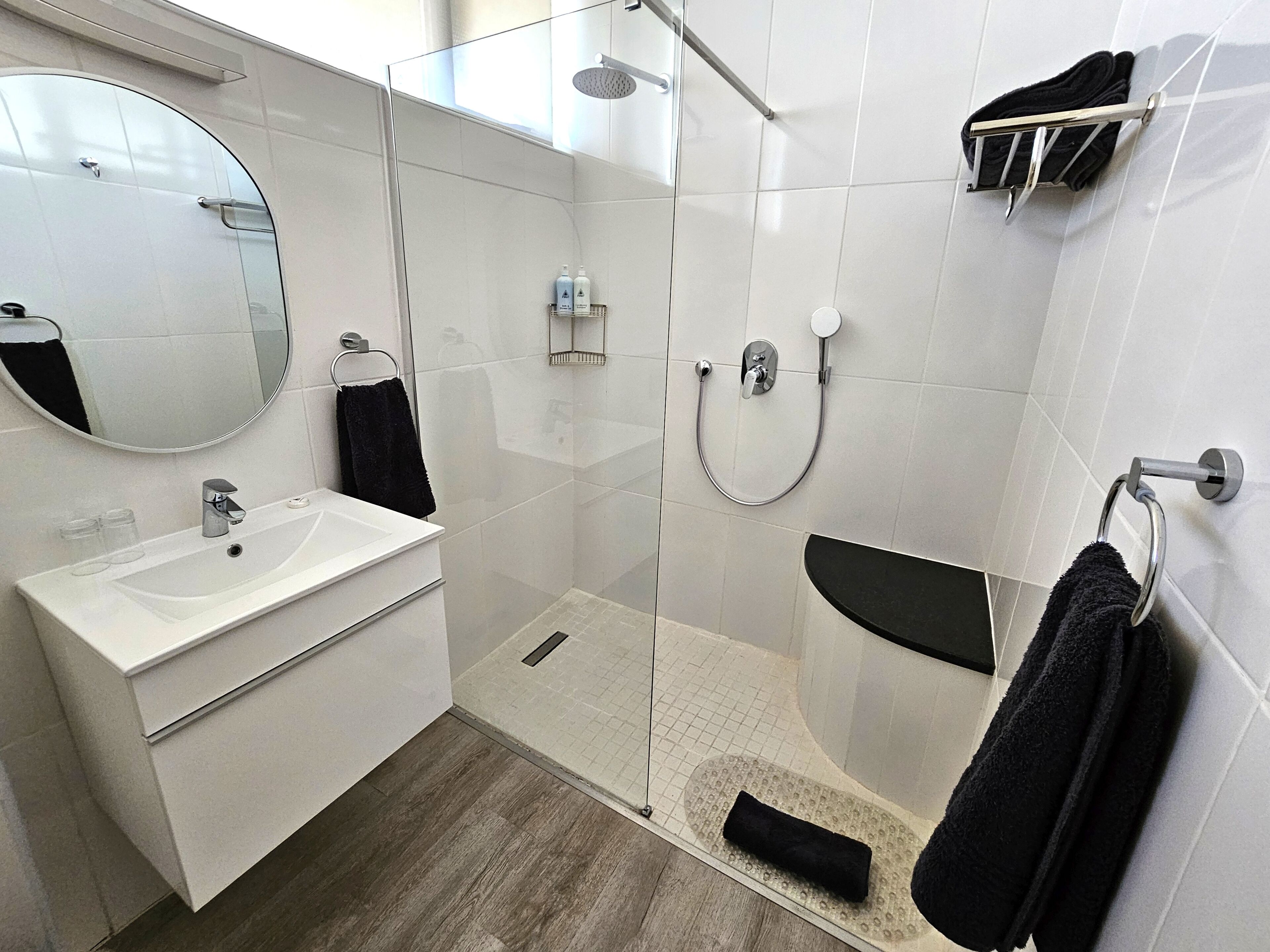 Un baño moderno con ducha envuelta en vidrio, un lavamanos blanco y un toallero negro.