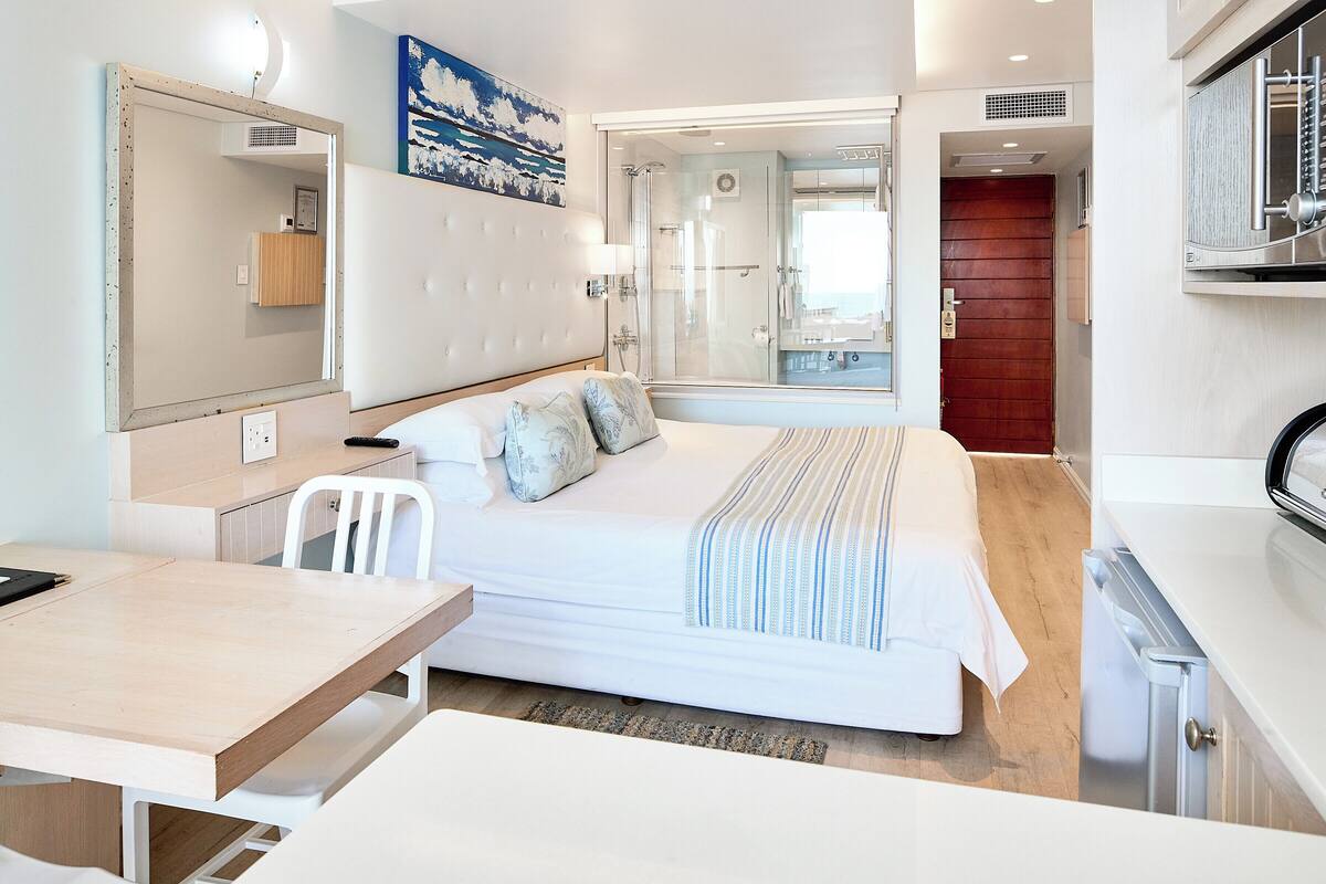 Una habitación de hotel moderna con cama, escritorio, silla, espejo y zona de ducha.