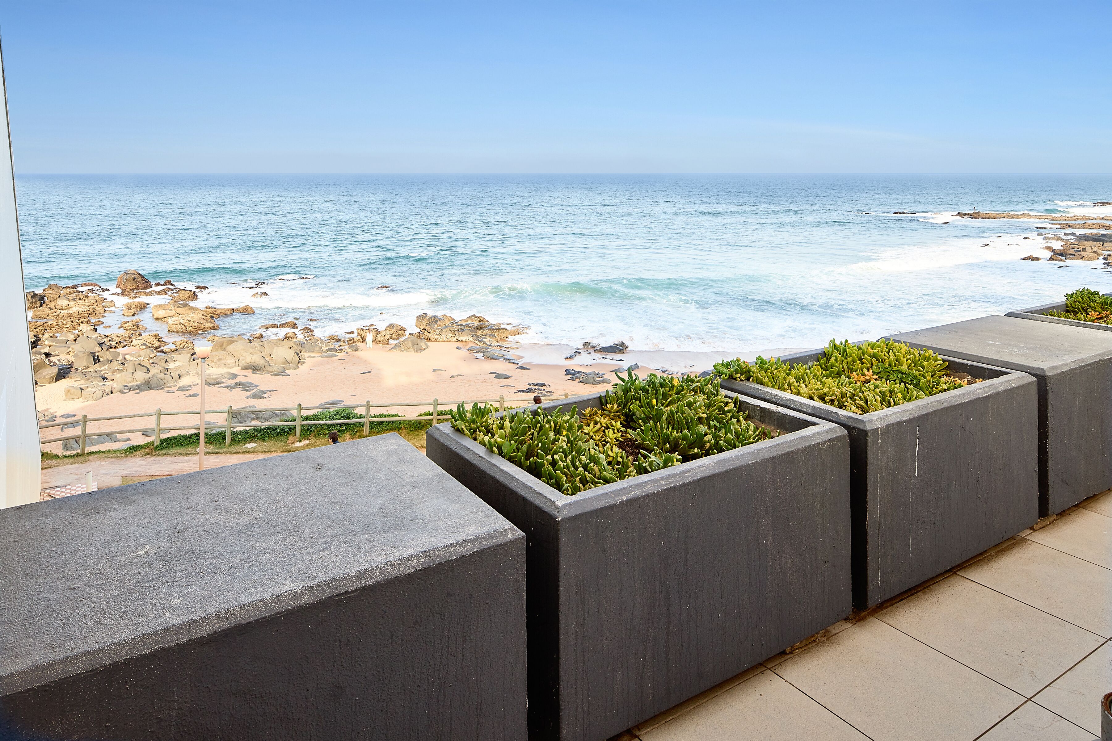 Un balcón con vistas al océano, macetas de concreto con plantas y una playa de arena con rocas.