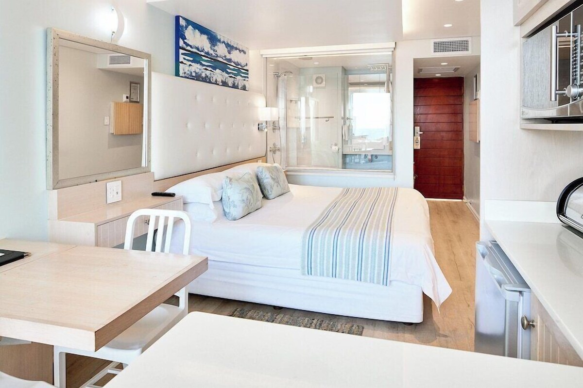 A modern hotel room with a bed, a desk, a chair, a mirror, and a shower area.
