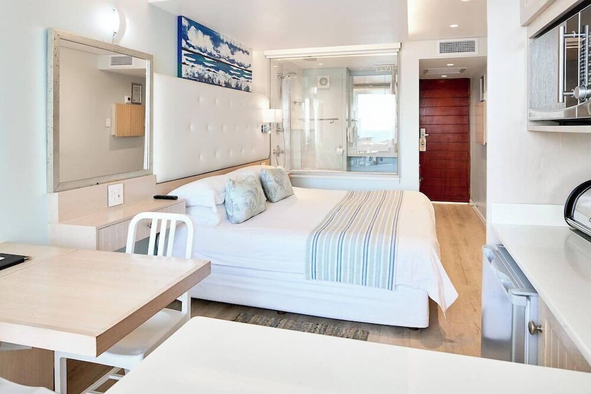 A modern hotel room with a bed, a desk, a chair, a mirror, and a shower area.
