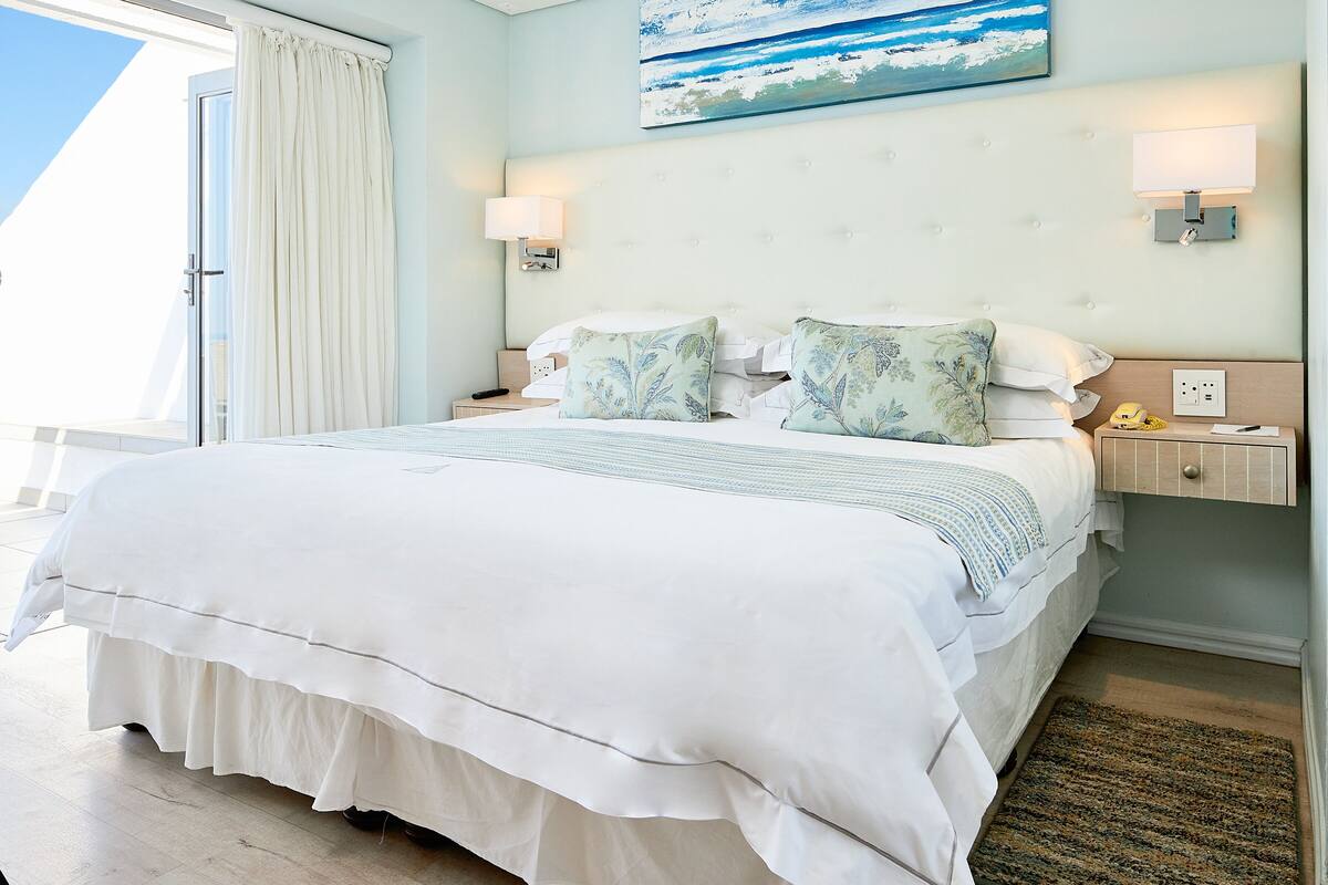 Habitación de hotel con una cama grande, dos mesitas de noche, un atril, un cuadro con vista al océano y vistas al exterior.
