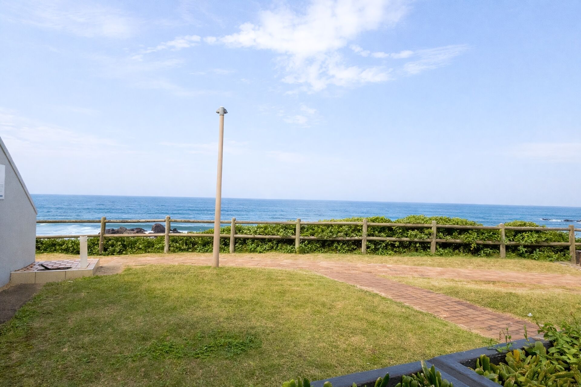 A grassy yard with a wooden fence, a paved path, and a view of the ocean.