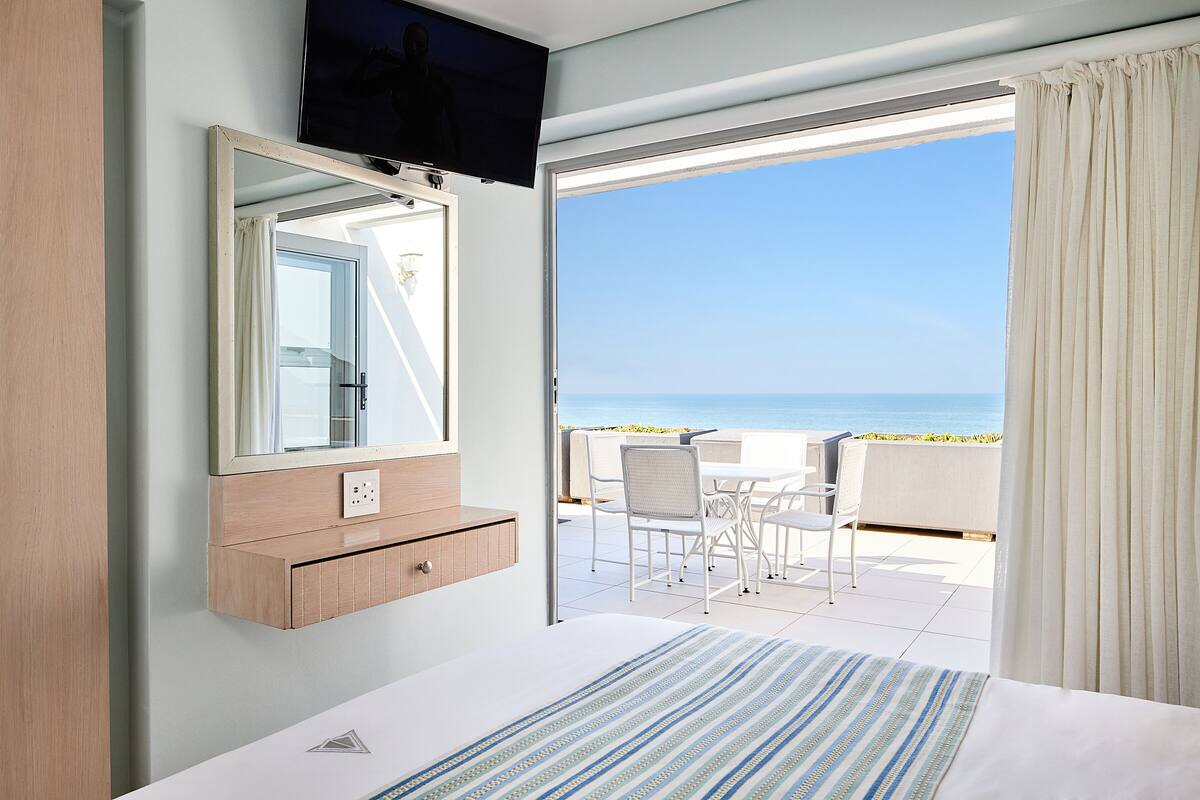 Habitación de hotel con cama, televisor montado en la pared, baño con espejo y balcón con mesa y sillas con vistas al mar.
