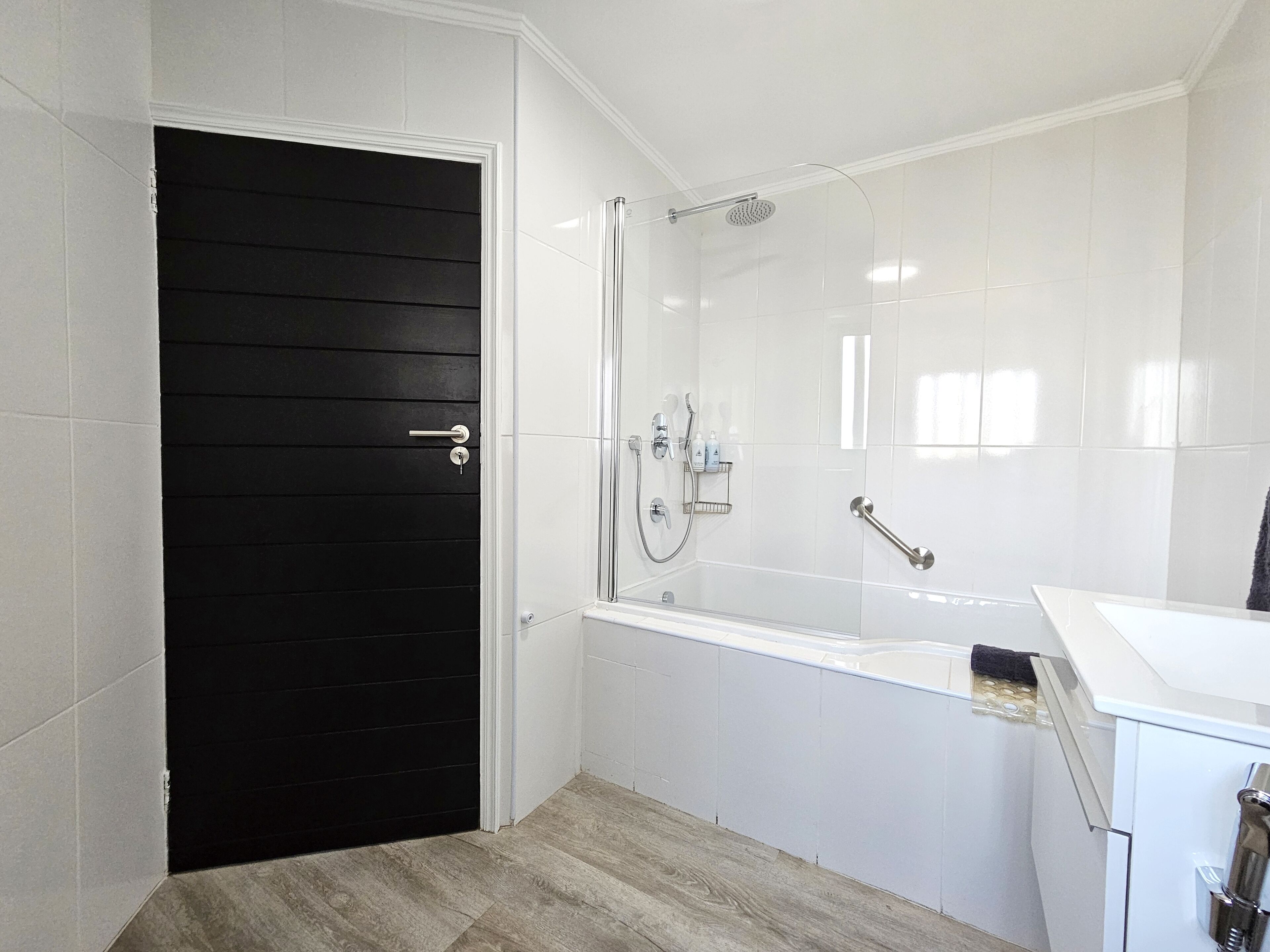 A modern bathroom with a black door, white tiled walls, a glass-enclosed shower, and a white sink.