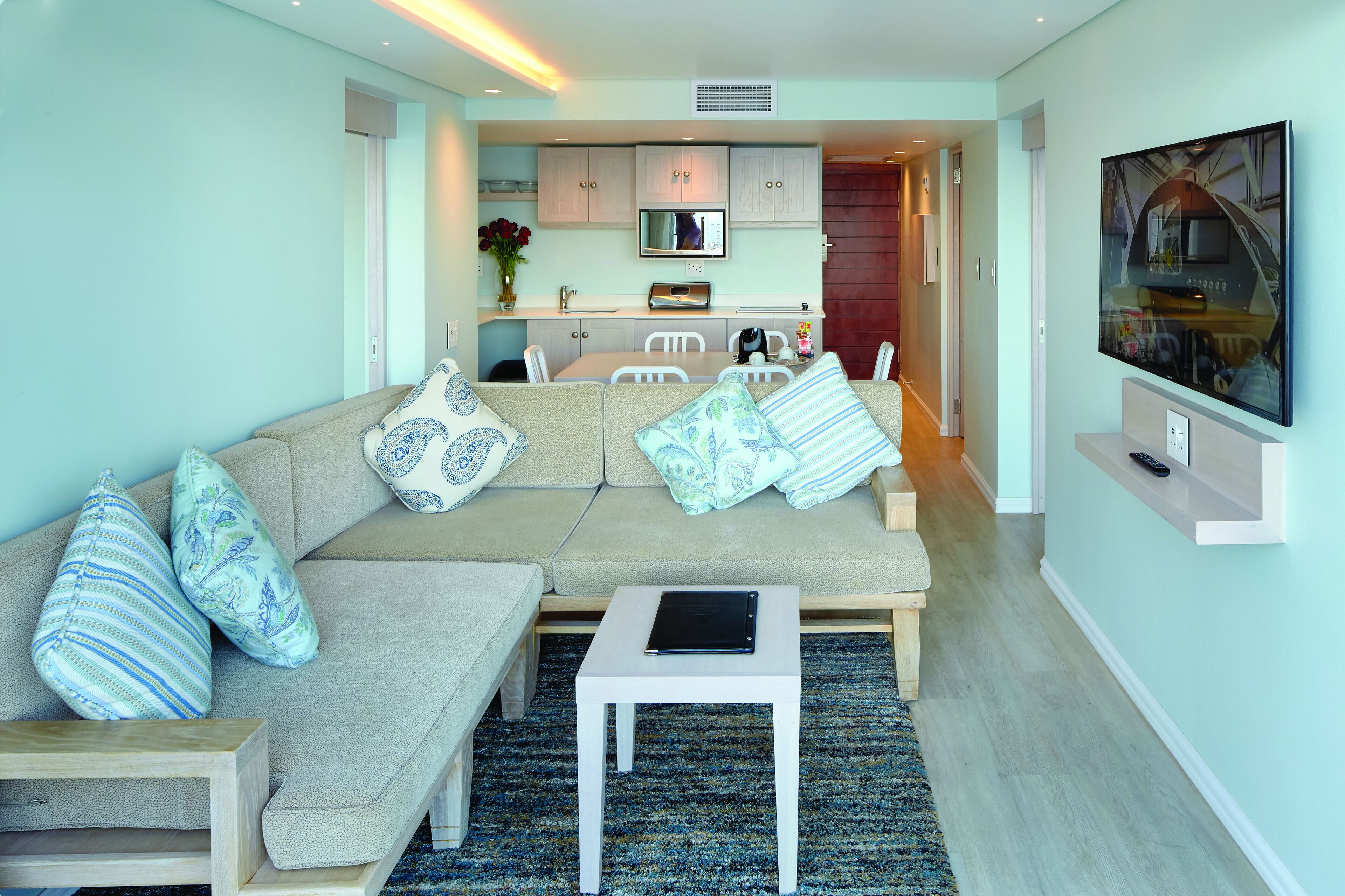 Ein modernes Wohnzimmer mit einem beigen Sofa, gemusterten Kissen, einem kleinen weißen Tisch und einem flachbildfernseher an der Wand.