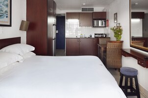 Una habitación de hotel con cama, kitchenette pequeño, espejo y silla de mimbre.