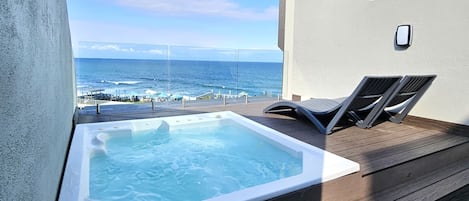 A balcony with a hot tub overlooking the ocean, wooden deck, and lounge chairs.