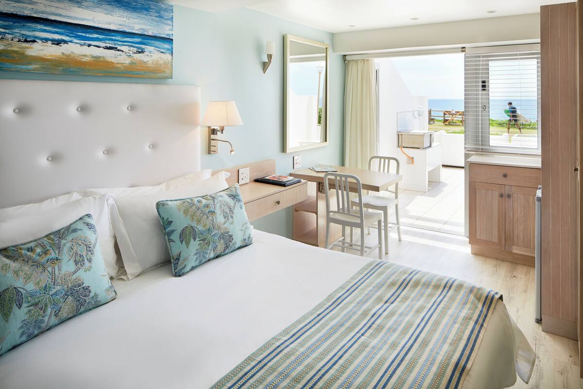 Habitación de hotel con cama, escritorio, silla, ventana grande y balcón con vista al mar.