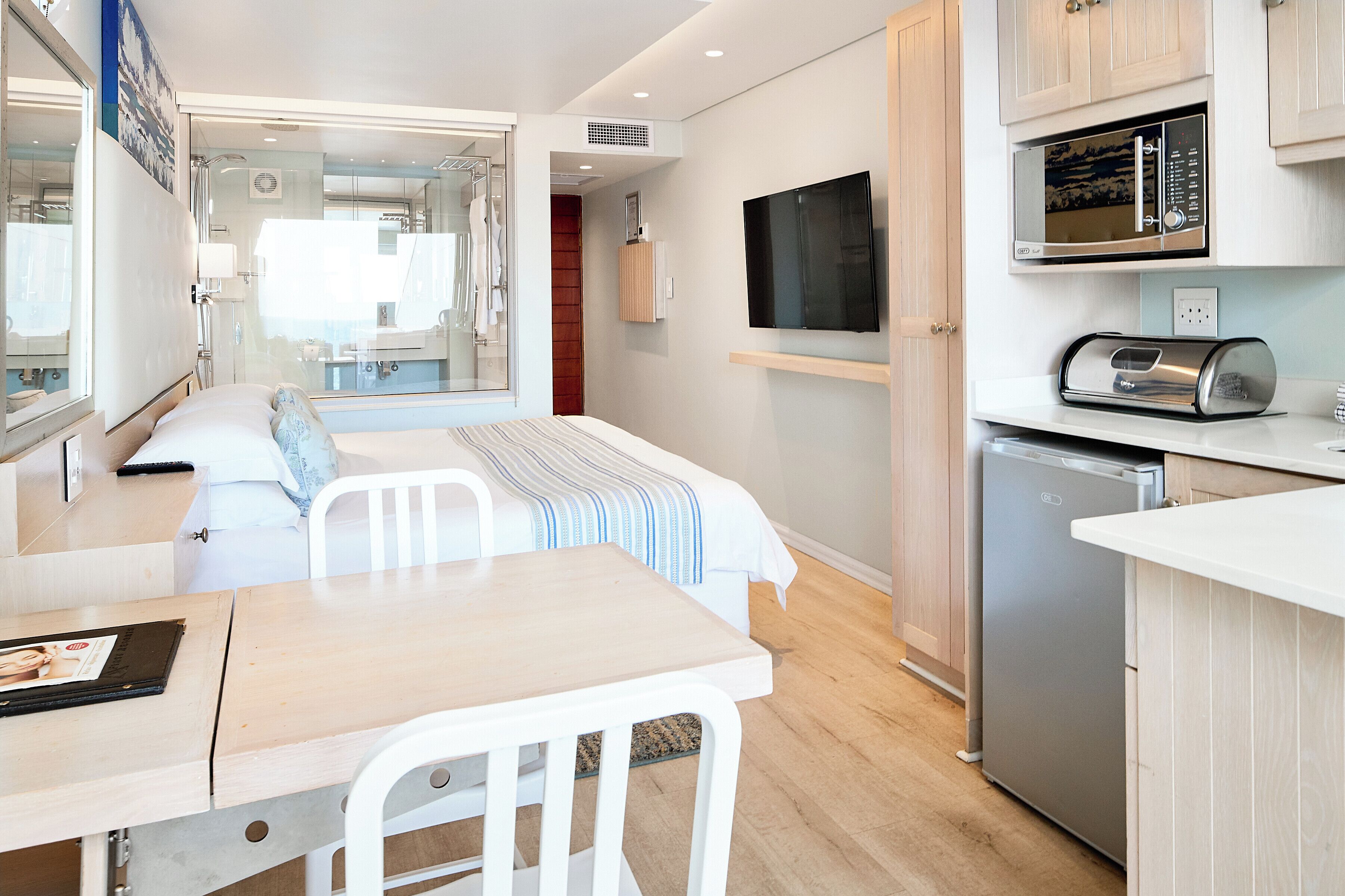 A compact hotel room with a kitchenette, a bed with a striped blanket, a TV mounted on the wall, and a window overlooking the interior.