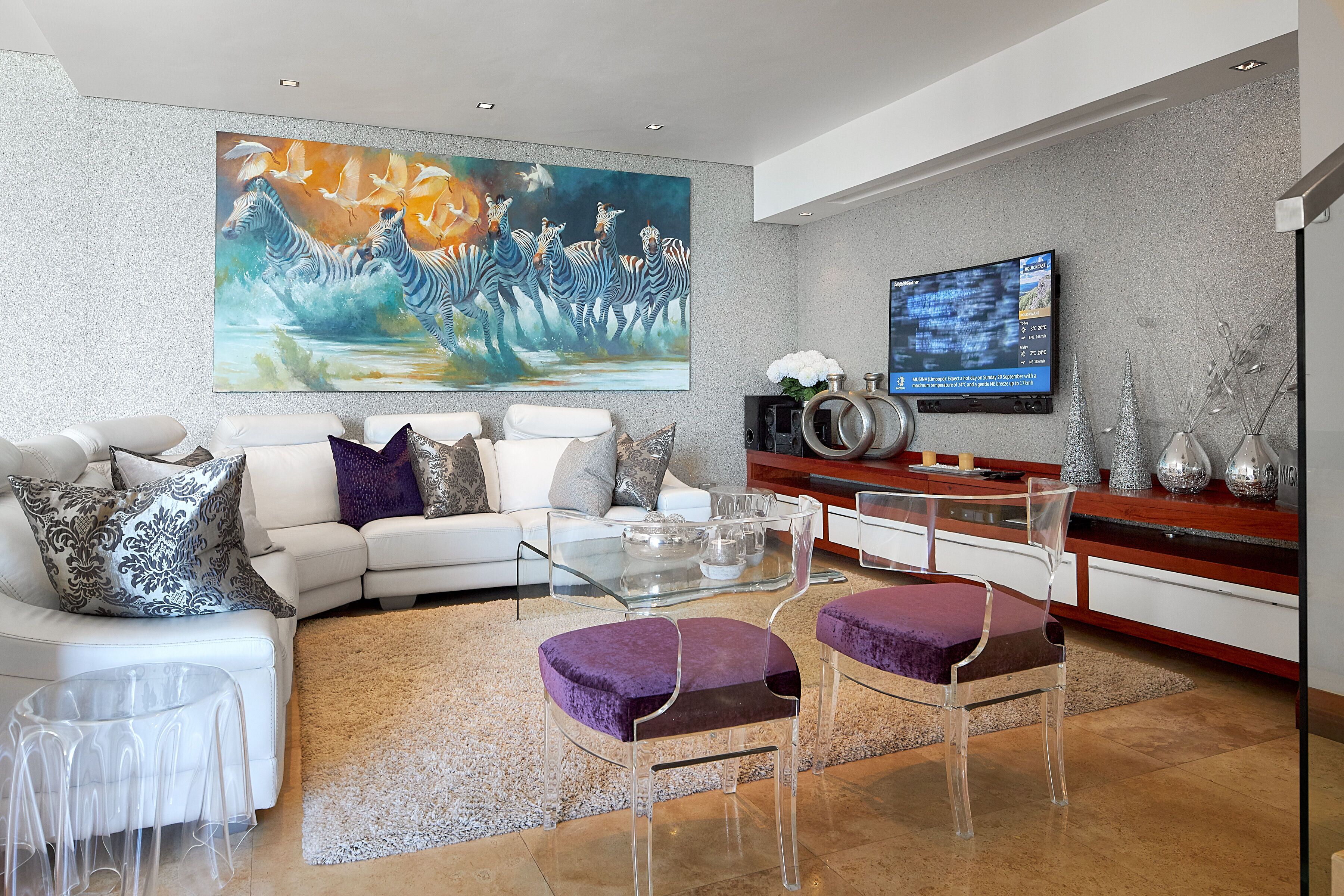 A modern living room with a white sofa, clear glass coffee table, and purple cushioned chairs. A large painting of zebras and birds is on the wall, and a TV is mounted above a wooden cabinet.