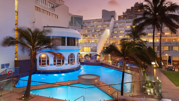Hotel com uma piscina grande, palmeiras e um edifício moderno com varandas.