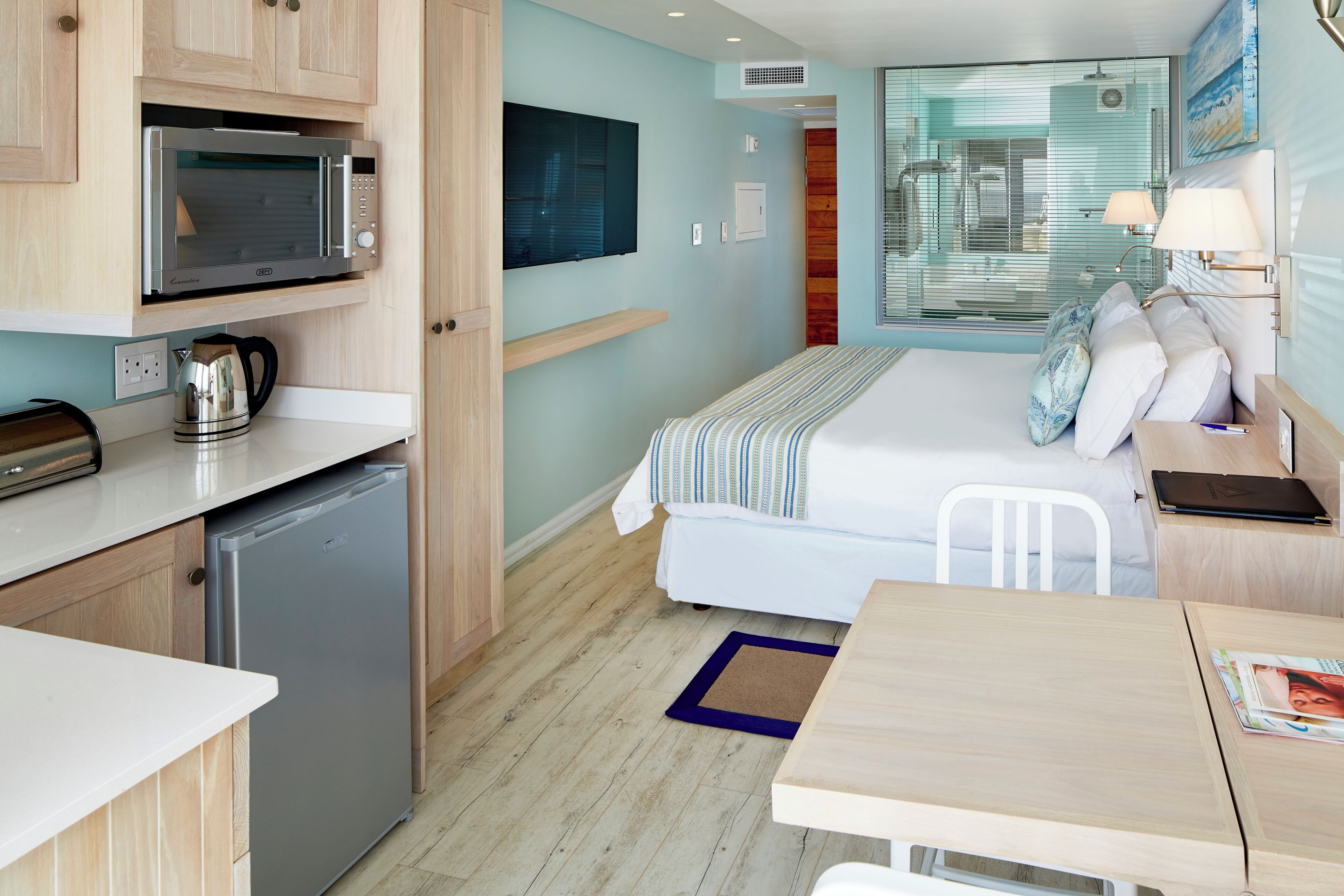 A compact hotel room with a kitchenette, a bed with a striped blanket, a bedside table, and a window with a view.