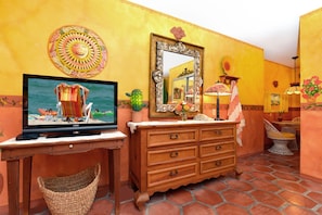 A room with a television on a wooden table, a wooden dresser with drawers, a mirror, and a wicker basket. - Blue Seas Courtyard (Lauderdale-by-the-Sea)