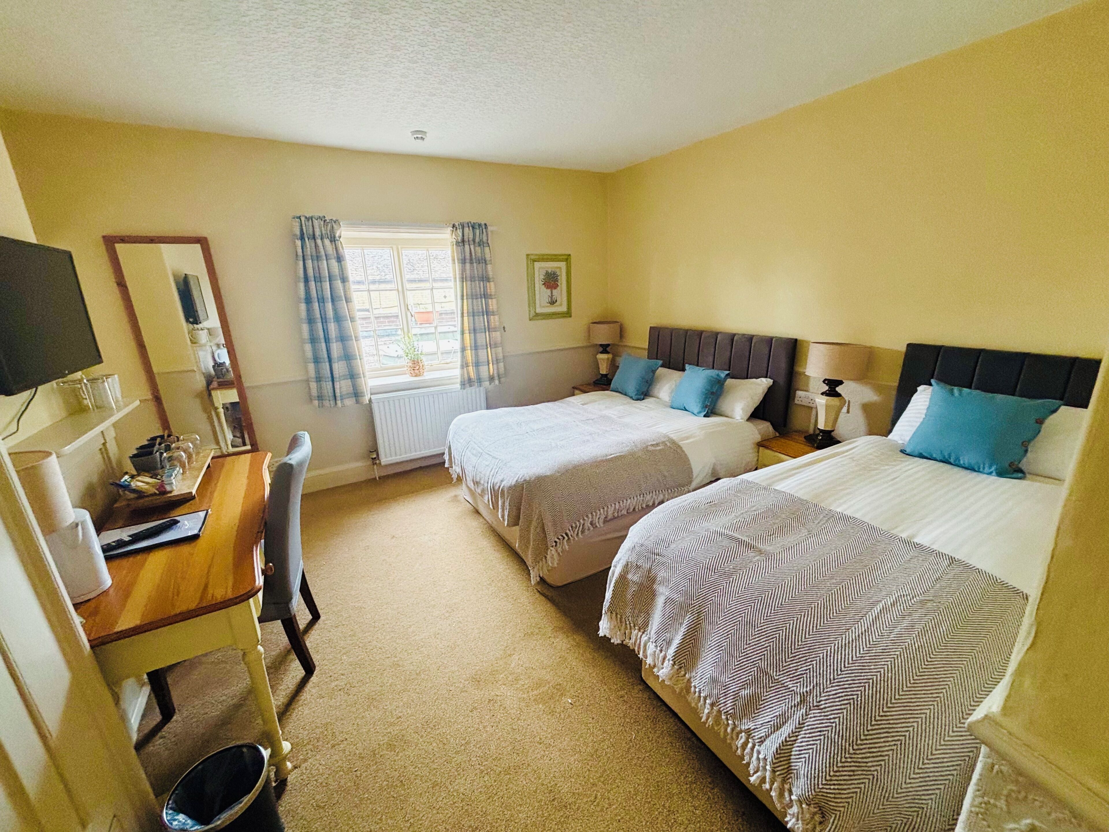 A hotel room with two beds, a desk with a chair, a window with curtains, and a television on the wall.